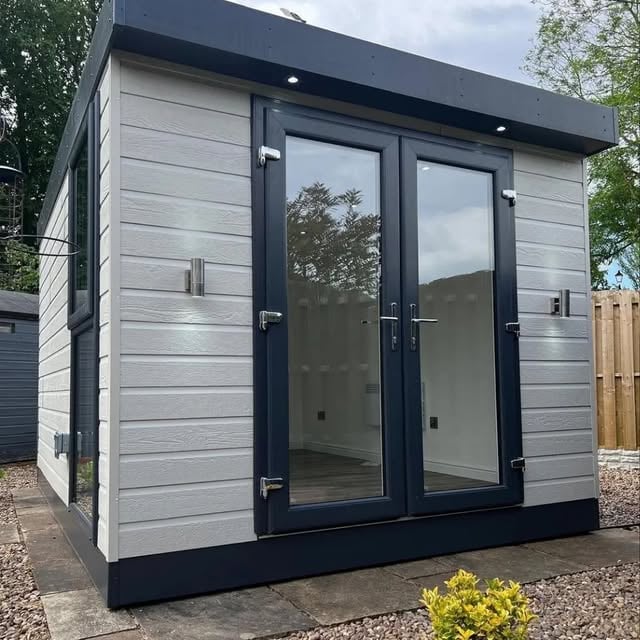 Modern garden studio with gray horizontal siding, black frame, glass double doors and black base on a gravel pad