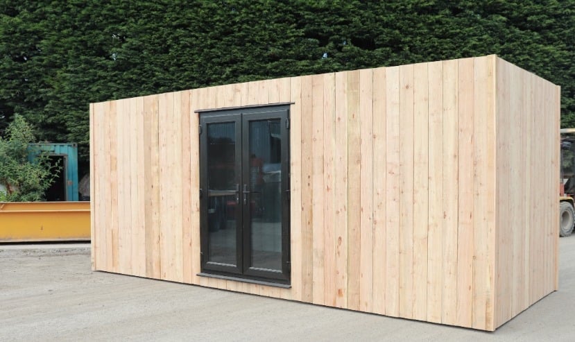 Modern wooden prefab cabin with glass door on a concrete patio with a green hedge in the background