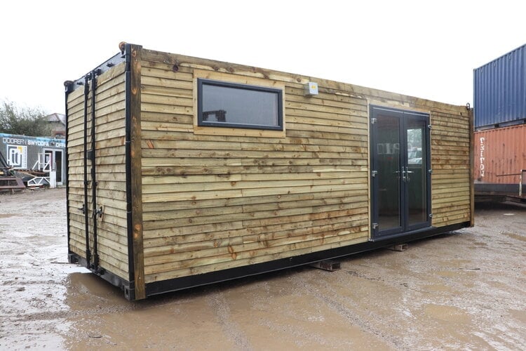 Wooden shipping container conversion with timber cladding, glazed double doors and a window, parked in an industrial yard