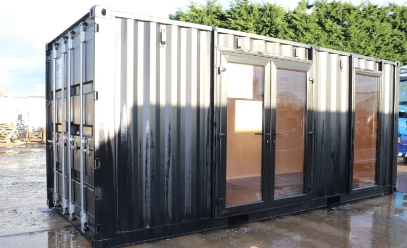 Modified shipping container with black corrugated metal exterior, glass doors and green plants on top in an outdoor lot